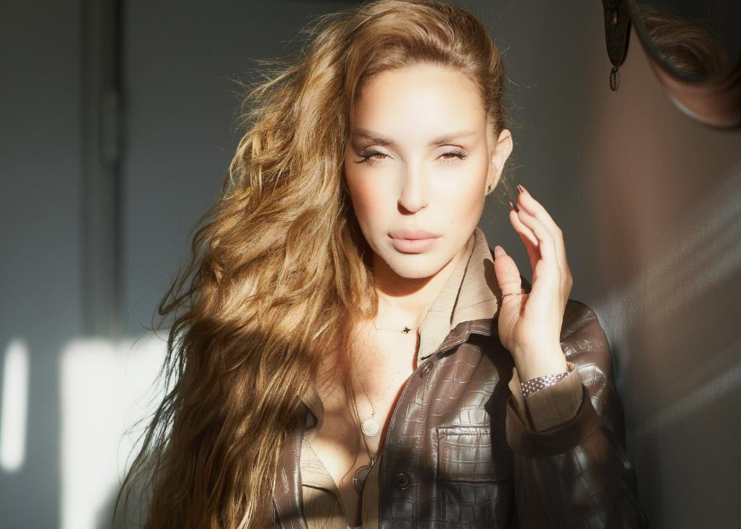 Woman with long brown hair wearing a brown leather jacket indoors.